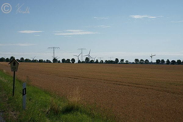 Blick über das LSG-Schild auf eine ausgeräumte Agrarlandschaft mit Windkraftanlagen